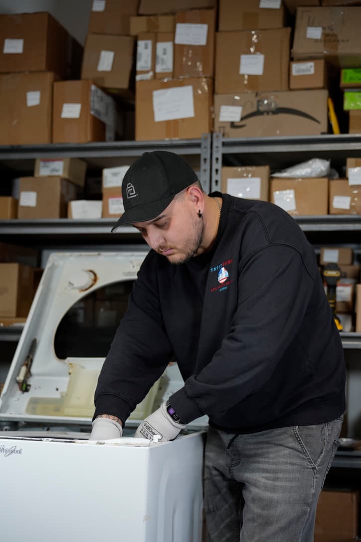 Trust Home technician in a black cap and branded black sweatshirt repairing a white top-loading washing machine