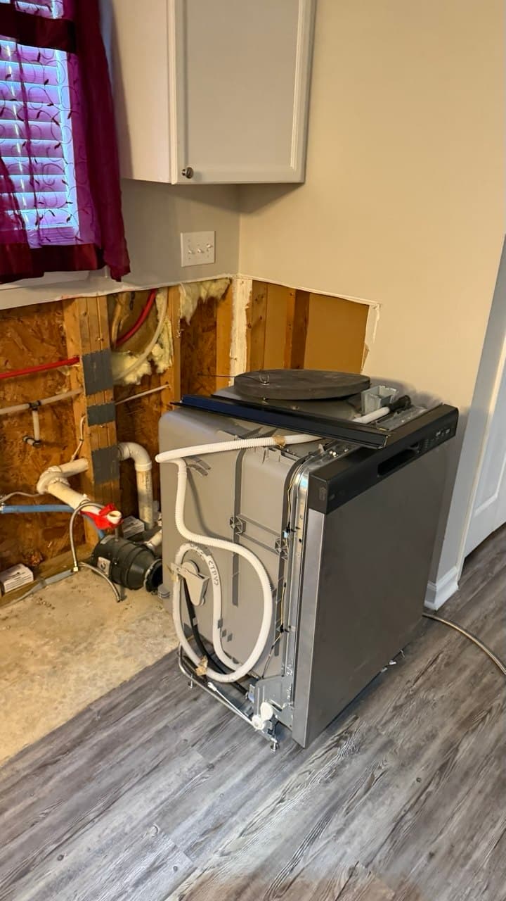 Stainless steel dishwasher pulled out from a kitchen wall, revealing exposed plumbing and wall studs.