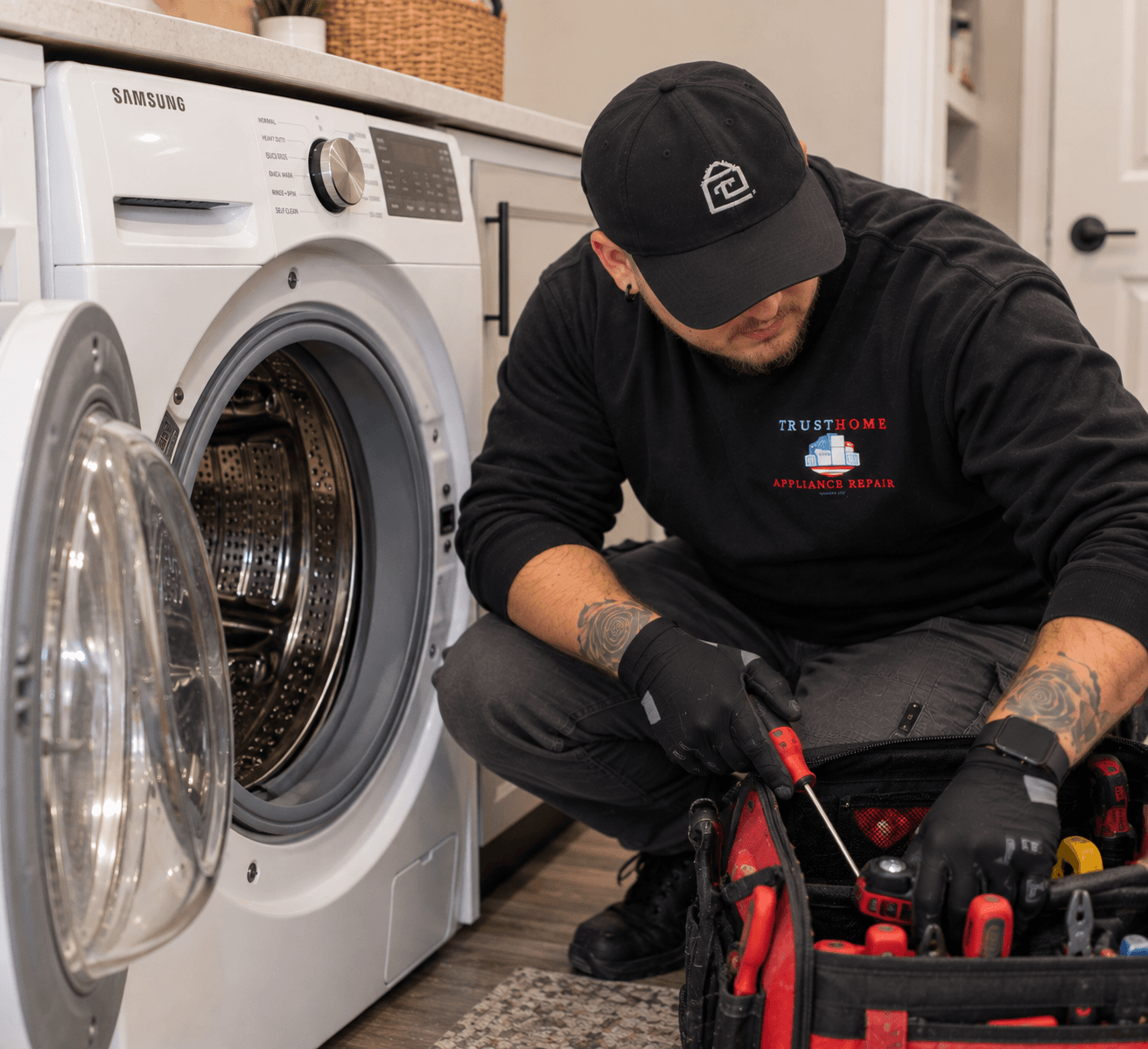 Appliance technician repairs a white Samsung front-load washing machine with tools from a bag.